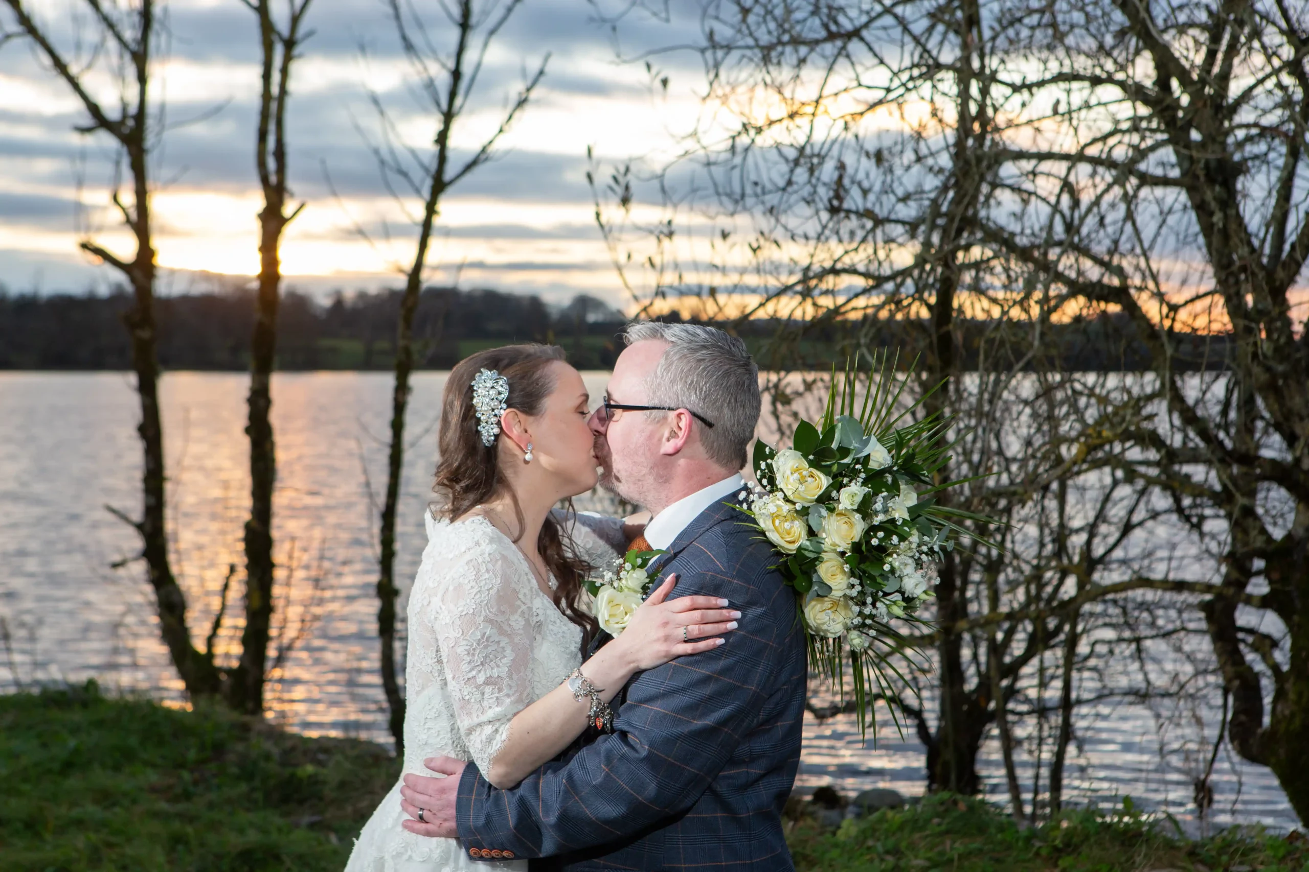 bride and groom stsnd kissing beside a lake and trees with a beautiful sunset sky