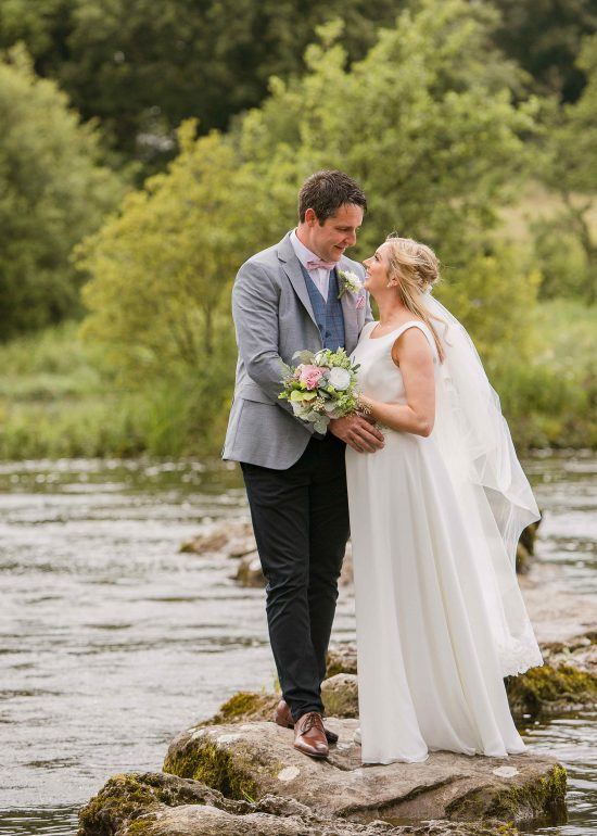 bride and groom stand on a stepping stone in a river