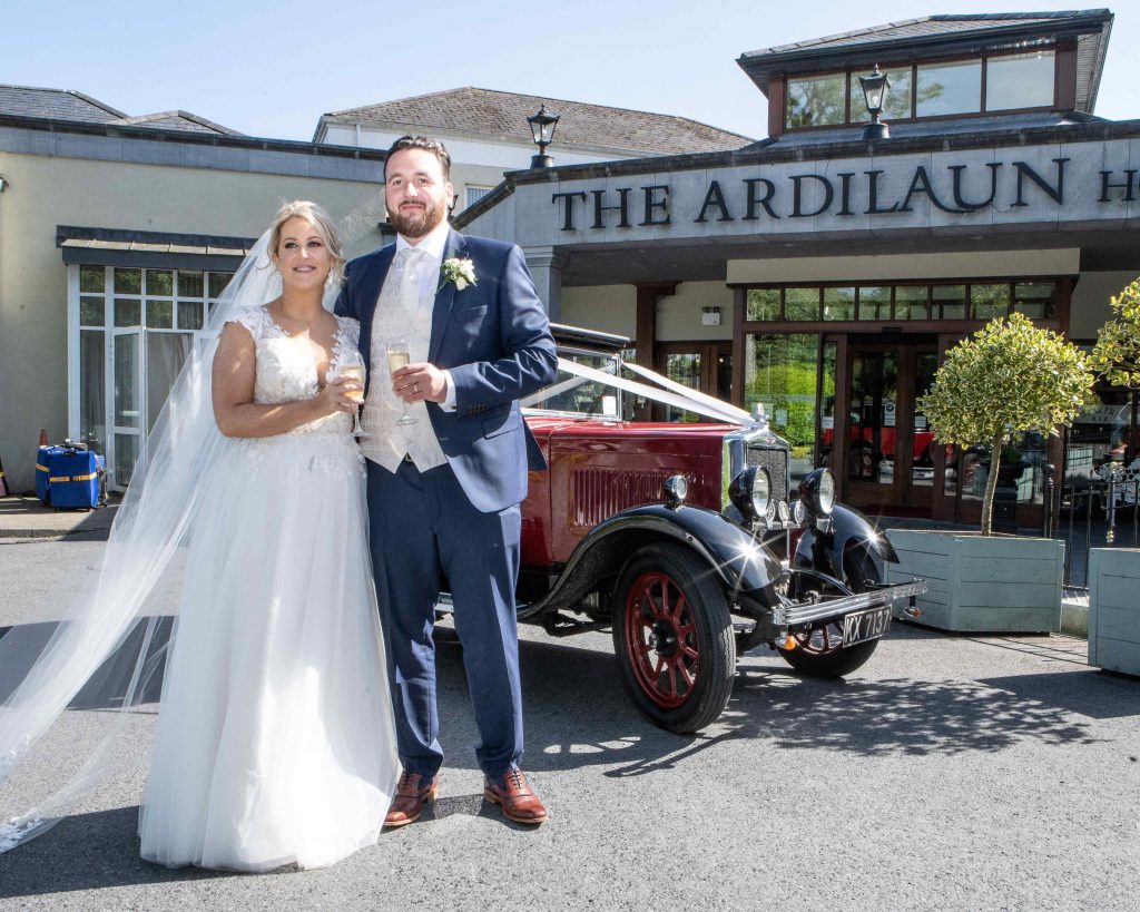 bride and groom stand in front of vintage red car outside the doors of Ardilaun Hotel Galway