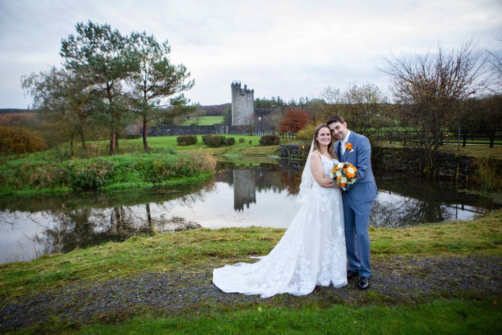 bride and groom stand beside a pond with a castle in the background
