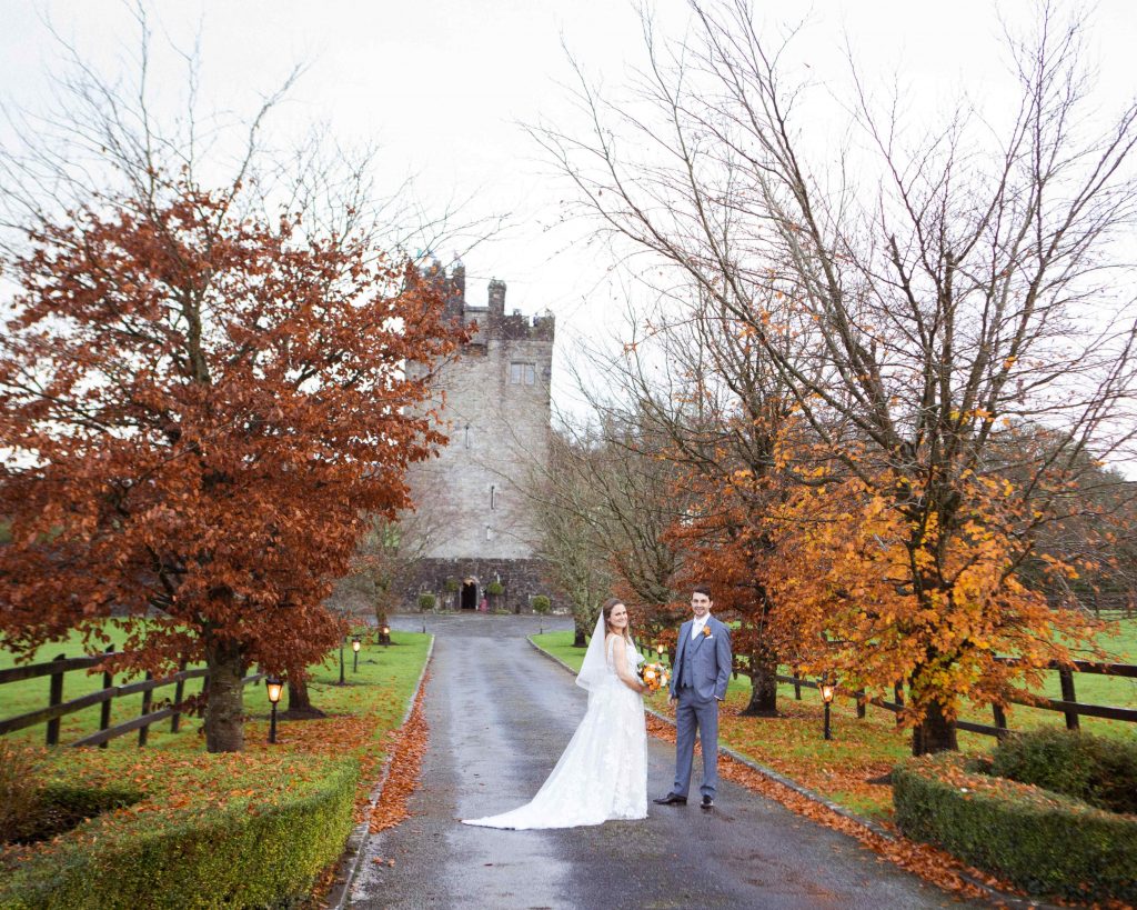 bride and groom stand on a treelined avenue with castle behind them