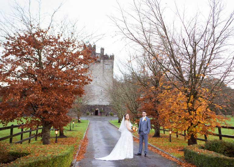bride and groom stand on a treelined avenue with castle behind them