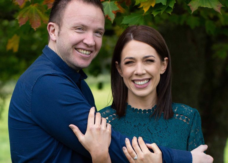 engaged couple stand under a tree red and green leaves, laughing