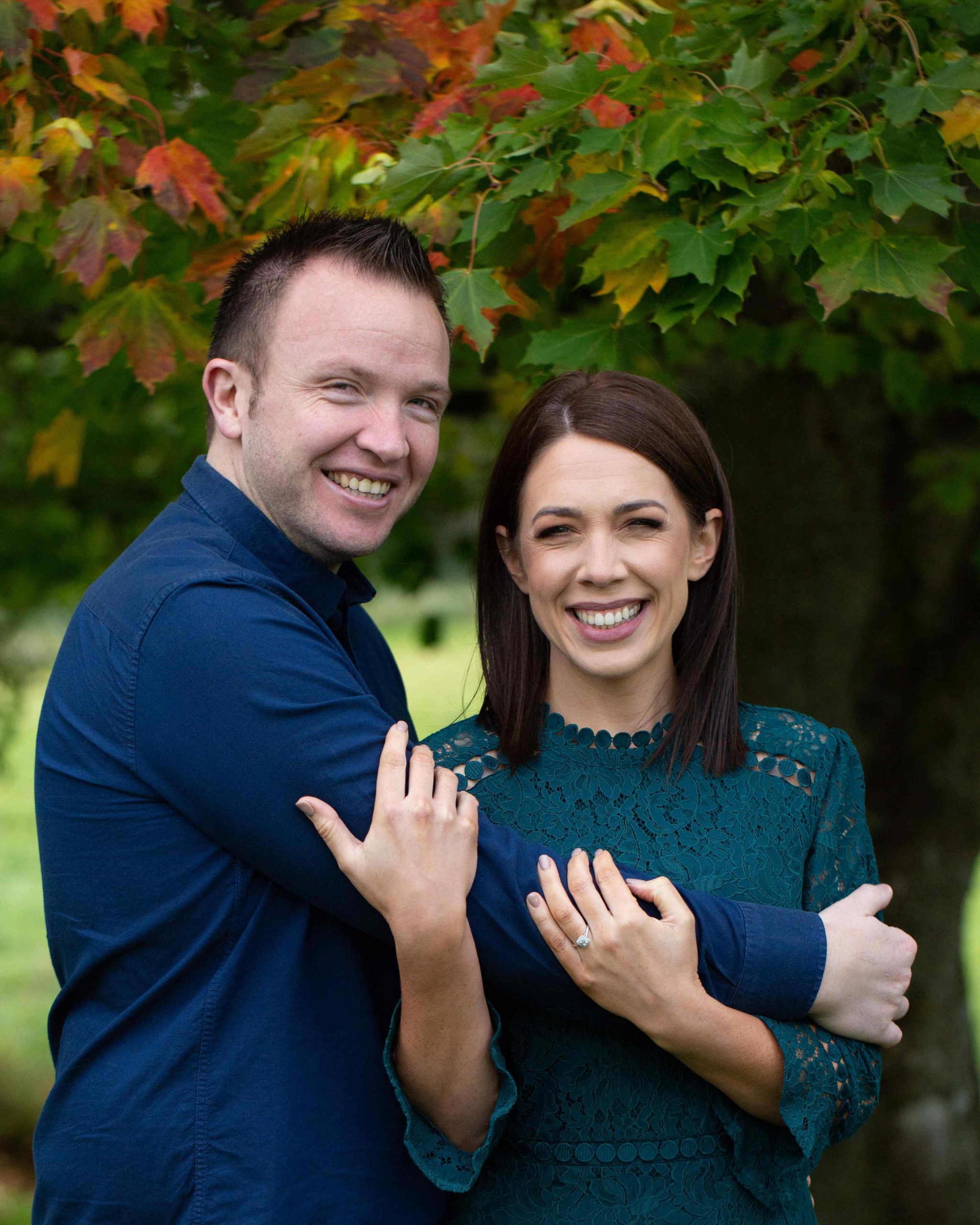 engaged couple stand under a tree red and green leaves, laughing