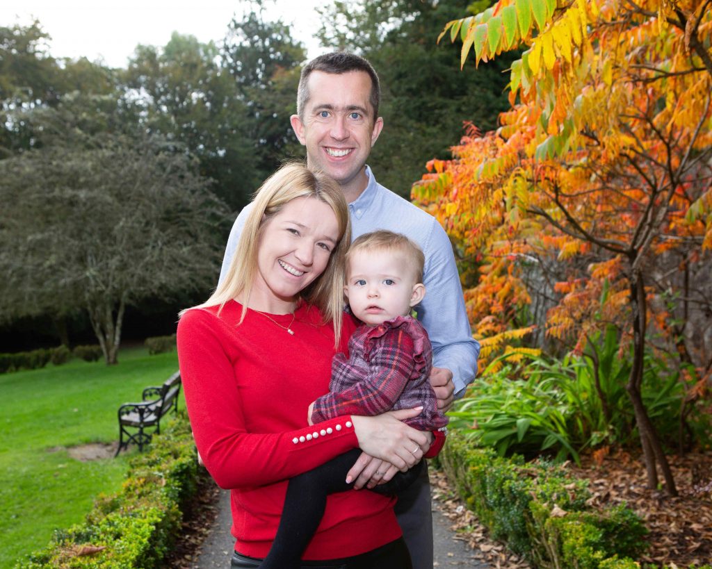 woman i red top holding toddler, man standing behind her , tree with orange leaves in the background