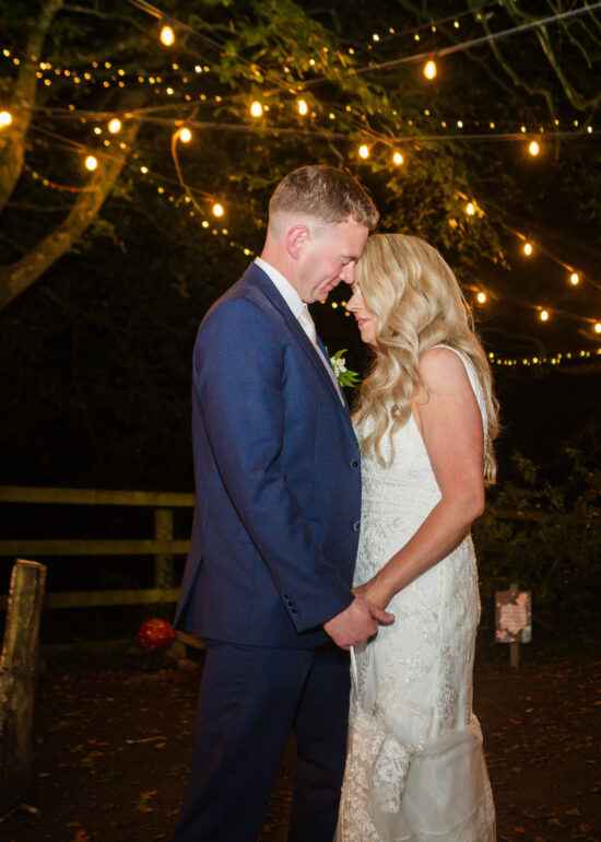 bride and groom standing under fairylights at night groom kissing top of brides head