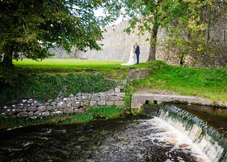 river and little weir in foregrond with bride and groom standing on the opposite bank under a tall tree