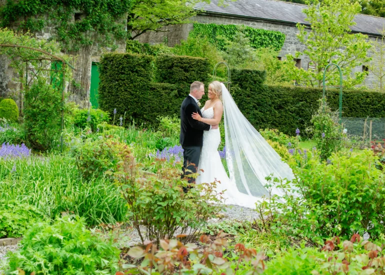 bride and groom stand embracing in a flowergarden with old stone building in the background