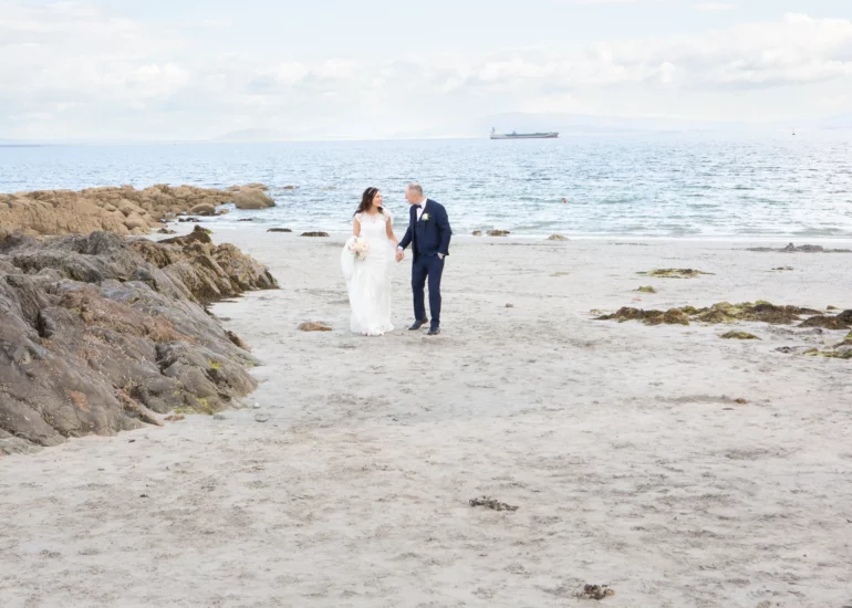 bride and groom walk holding hands on a sandy beach