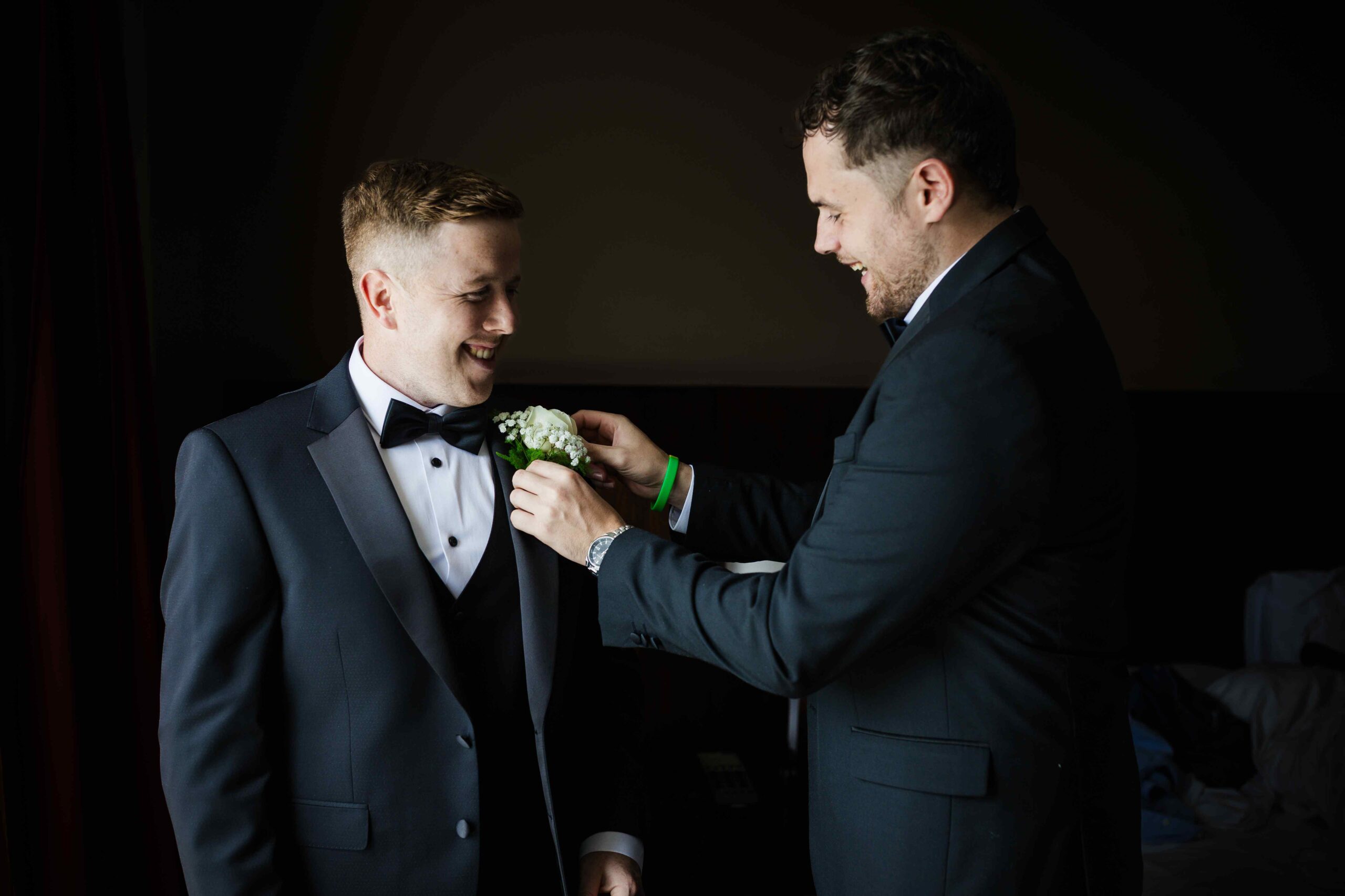 Groom smiling as groomsman fixes his buttonhole flower before the wedding ceremony.