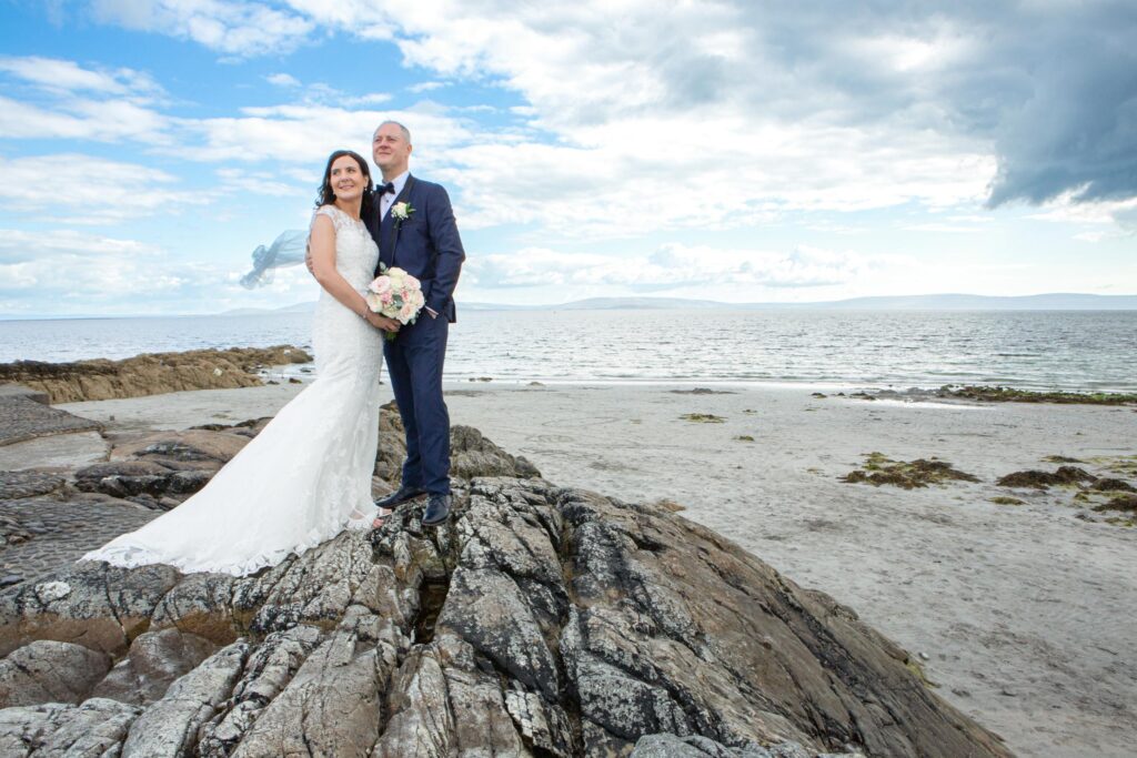 Bride and groom standing on rocks beside Galway Bay in Salthill
