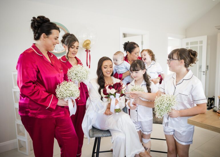 Bride seated with her bouquet, surrounded by bridesmaids and children during wedding morning preparations.
