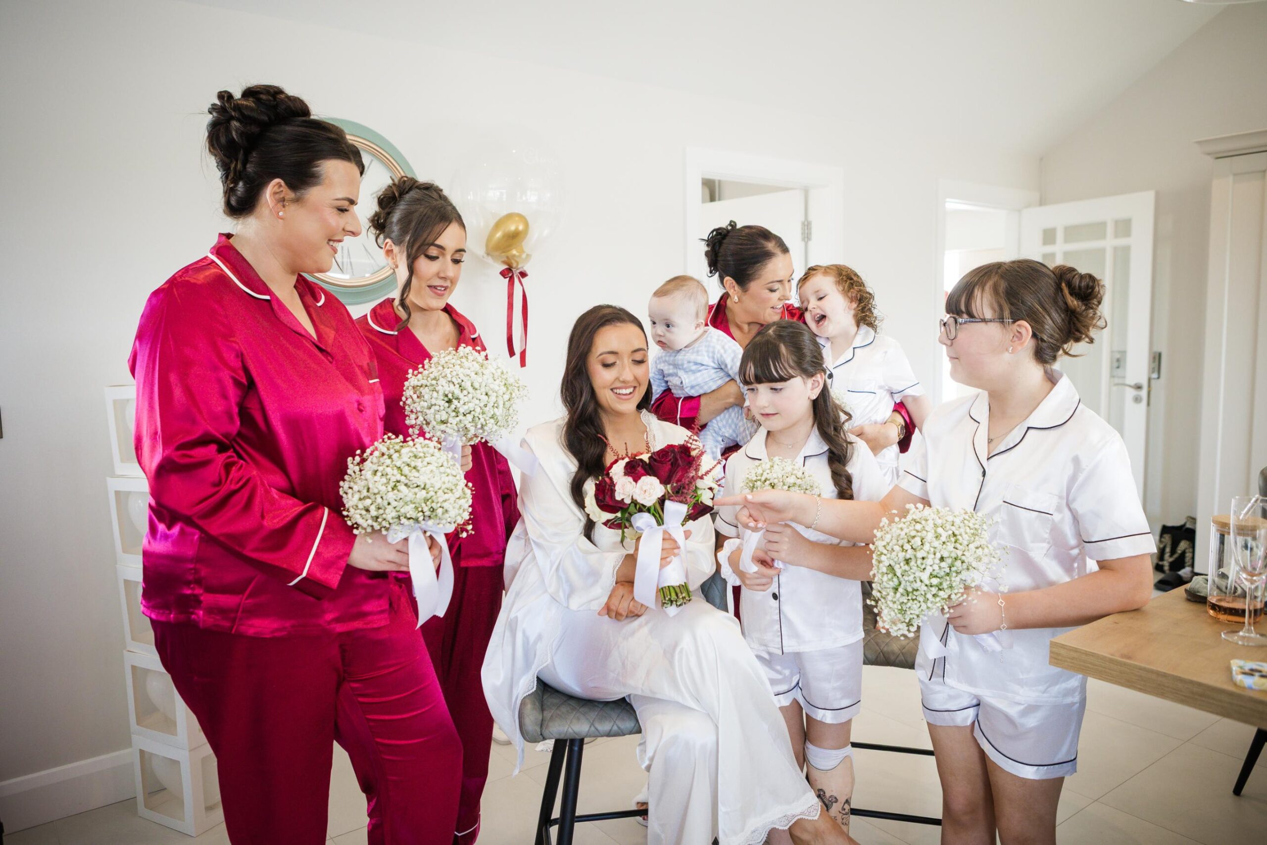 Bride seated with her bouquet, surrounded by bridesmaids and children during wedding morning preparations.