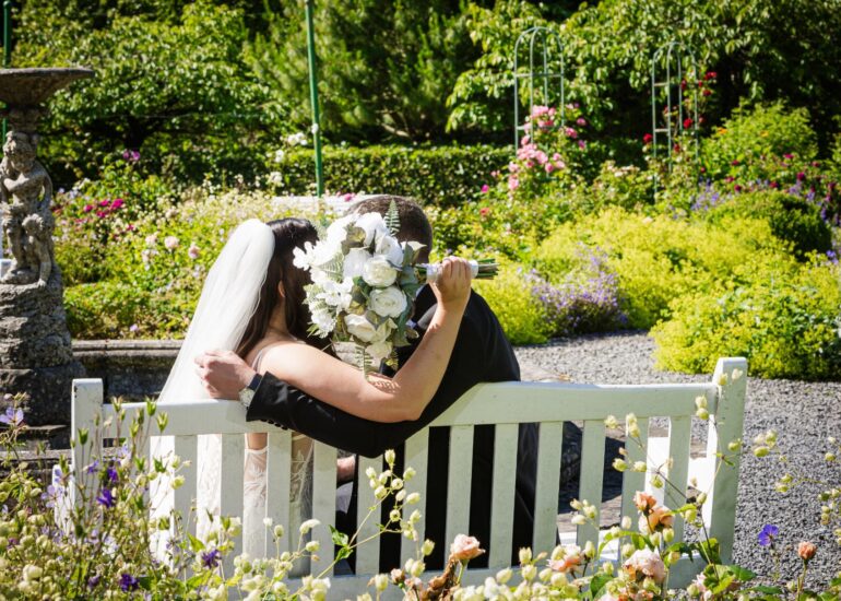 Bride and groom embrace on a garden bench, with the bride holding a white rose bouquet.