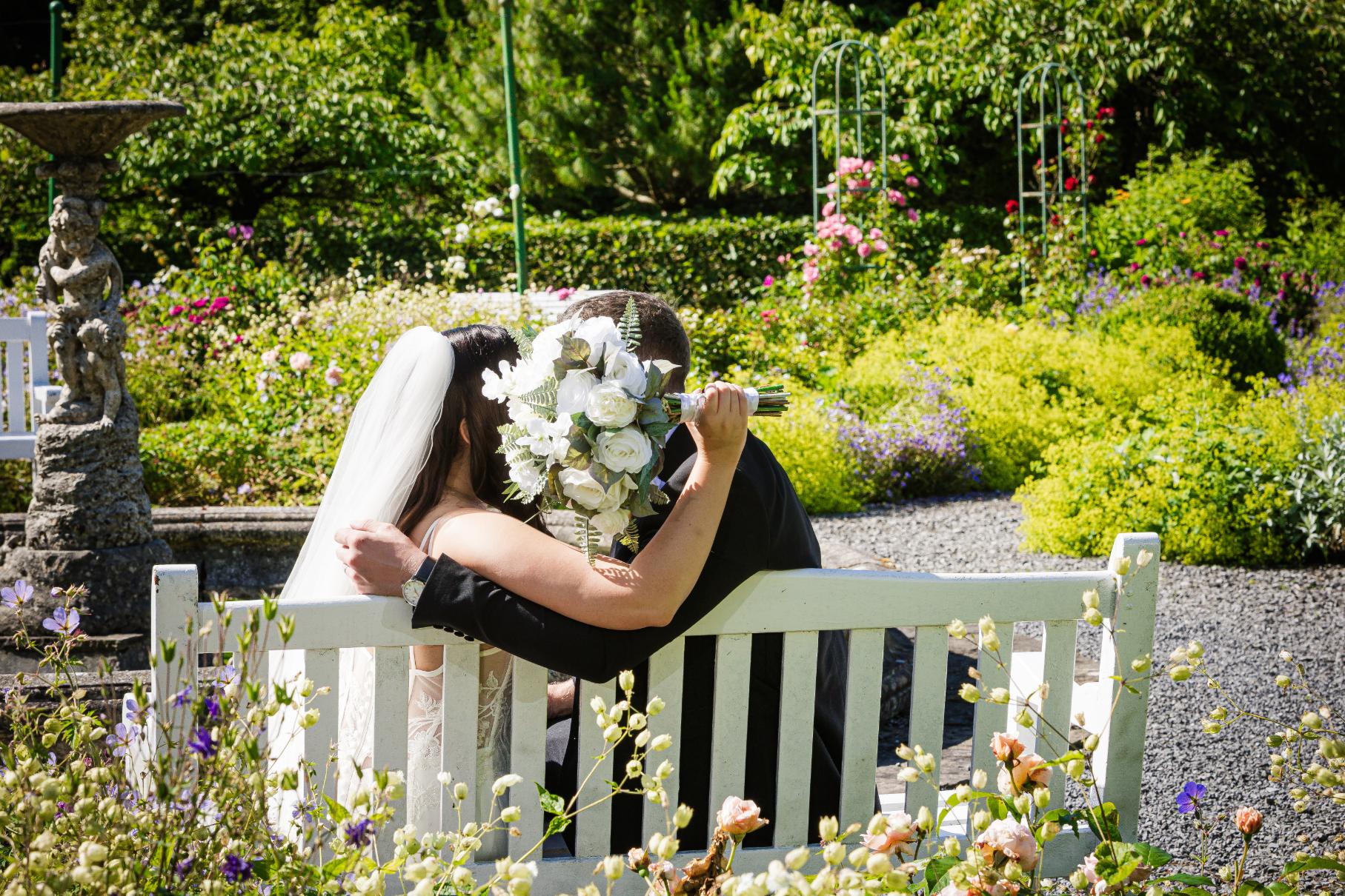 Bride and groom embrace on a garden bench, with the bride holding a white rose bouquet.