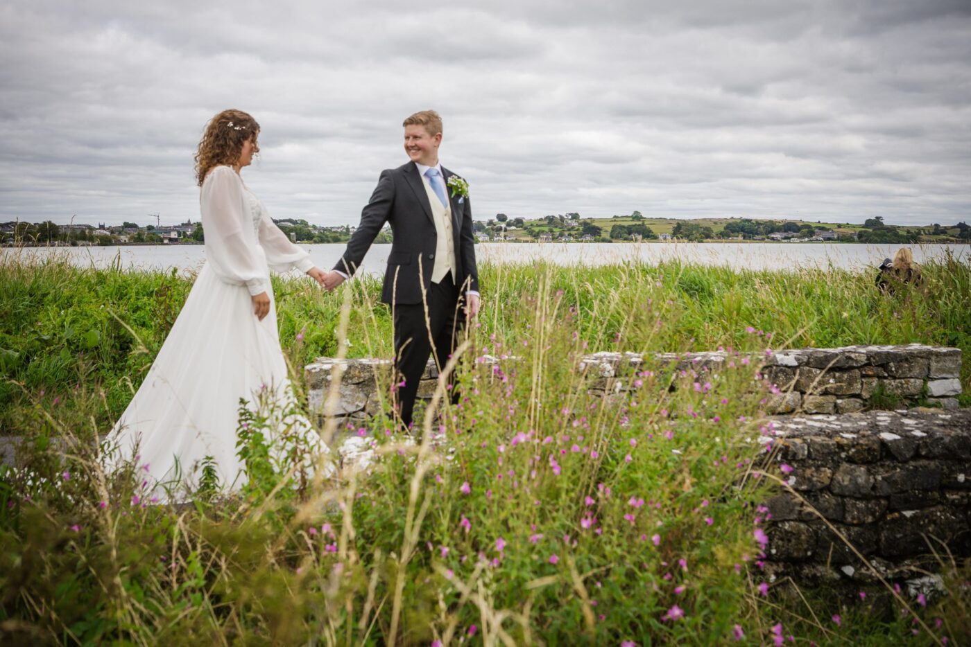 Bride and groom holding hands among tall grasses and pink wildflowers beside a lake.