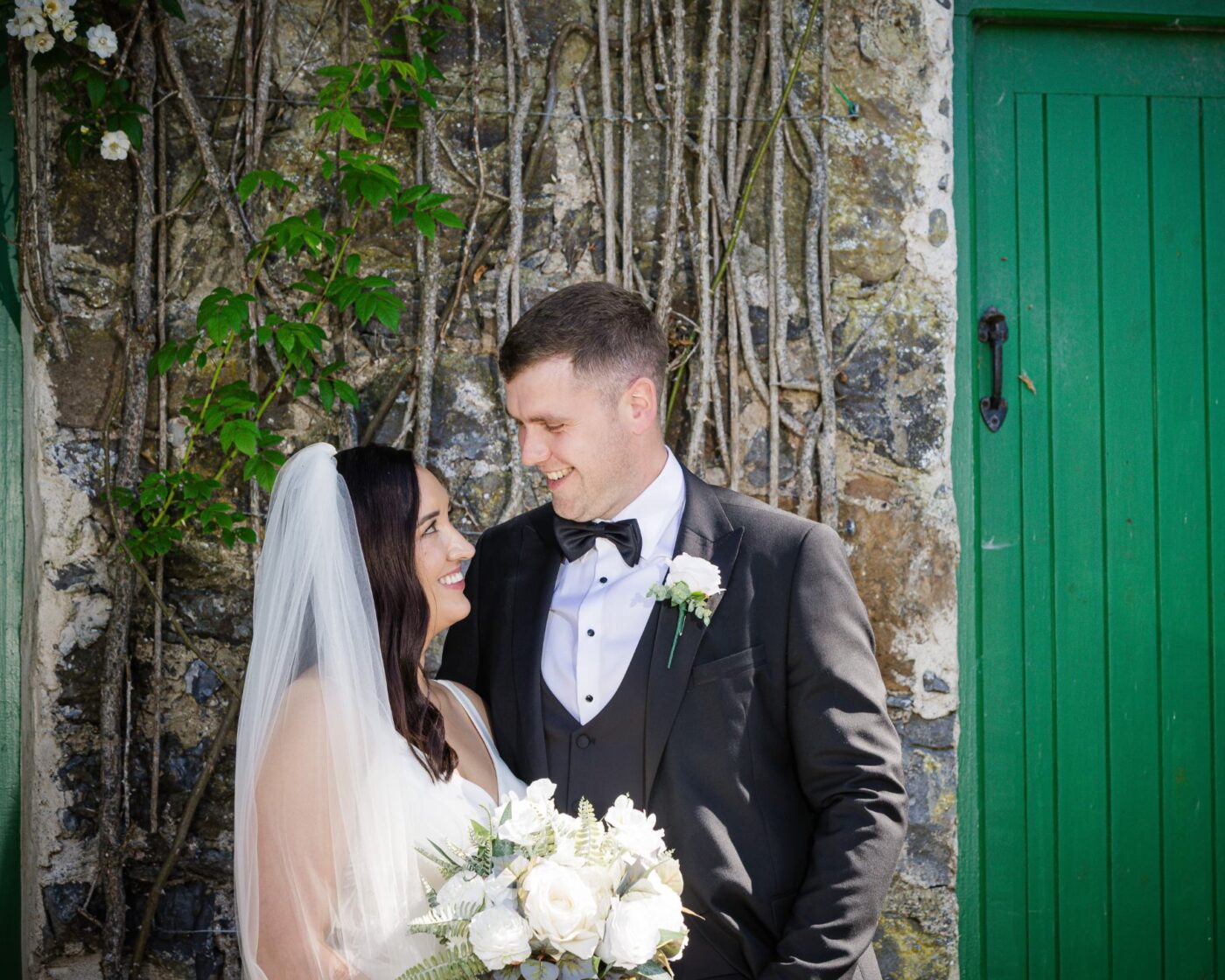 Bride and groom smiling at each other beside a green door and a climbing rose-covered stone wall.