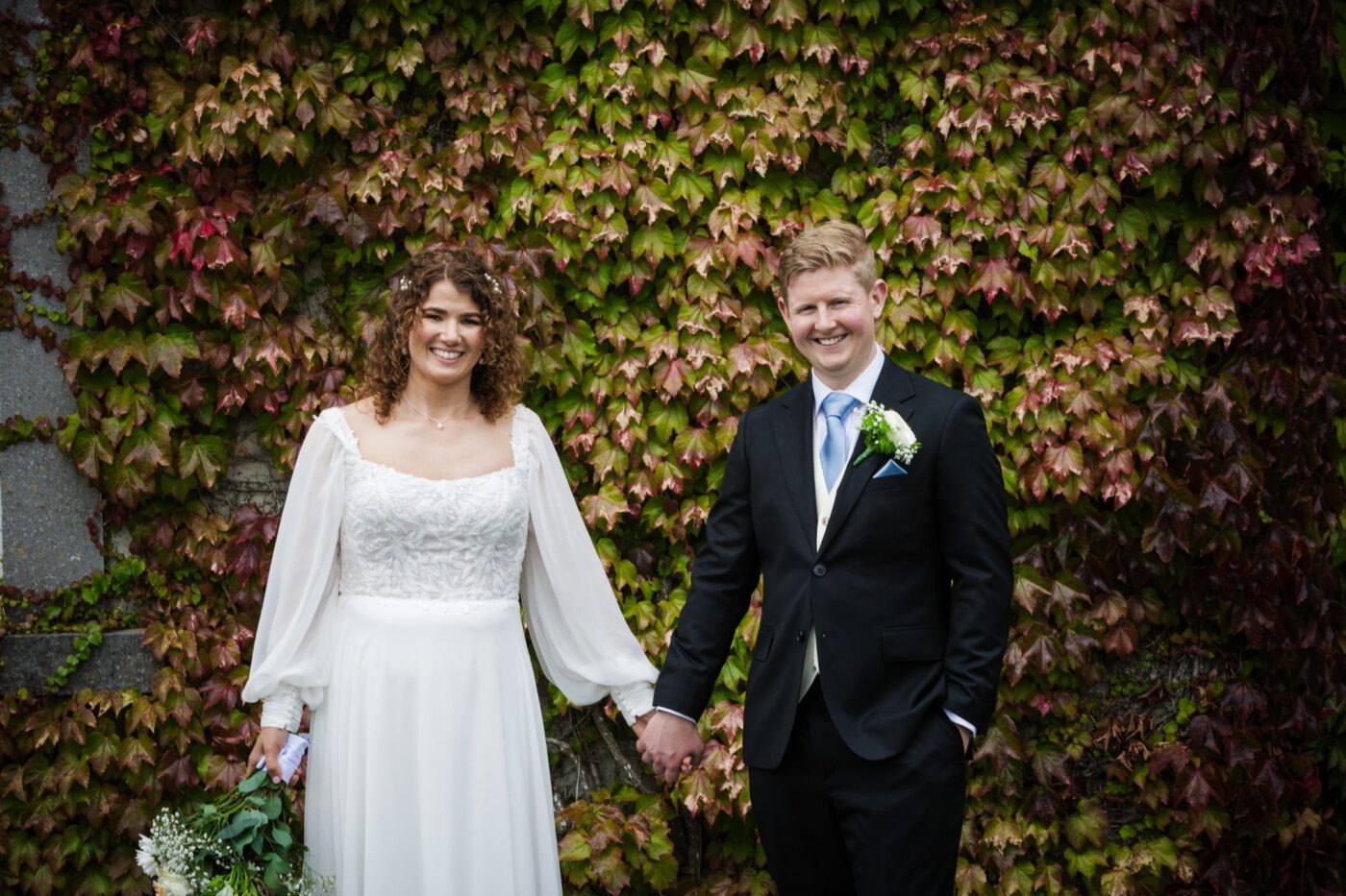 Bride and groom smiling and holding hands in front of an ivy-covered stone wall.