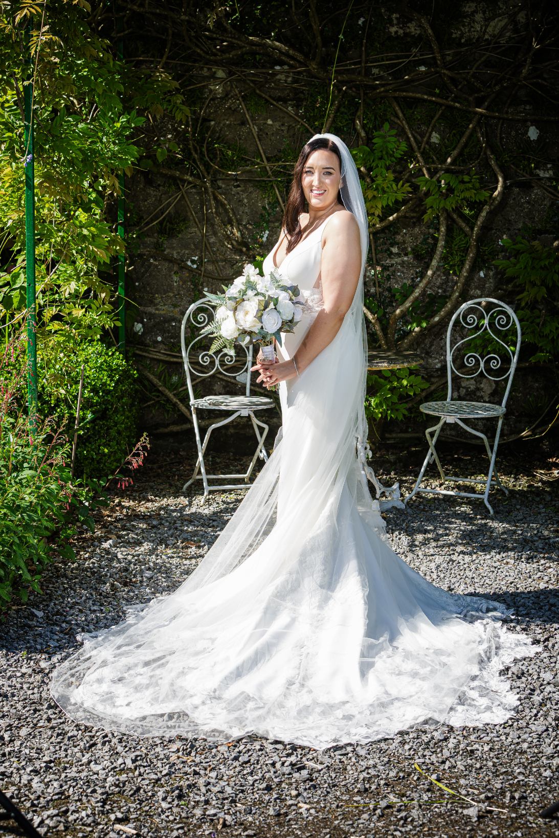 Bride holding a white rose bouquet in a sunlit garden, with her dress train spread across the gravel.