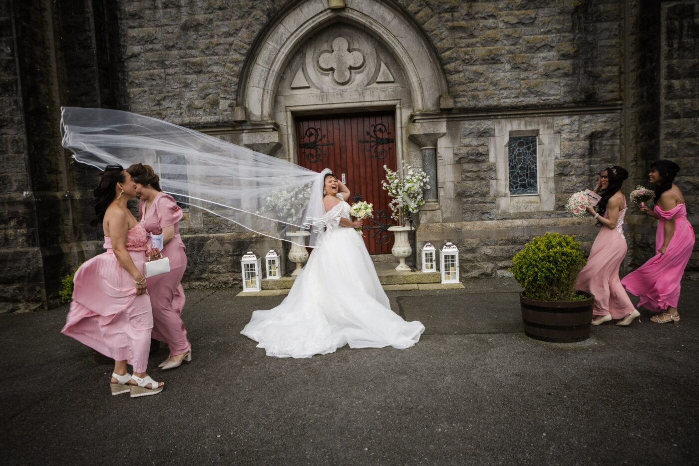 Laughing bride with attendants in pink outside a stone church as the wind lifts her long veil.