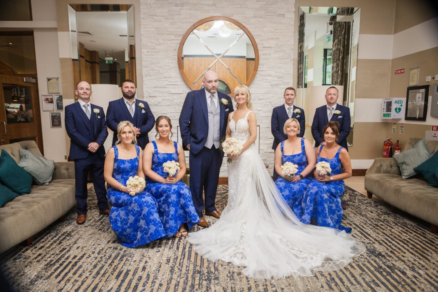 Bride and groom beside a hotel fireplace with seated bridesmaids in blue floral dresses and groomsmen in navy.