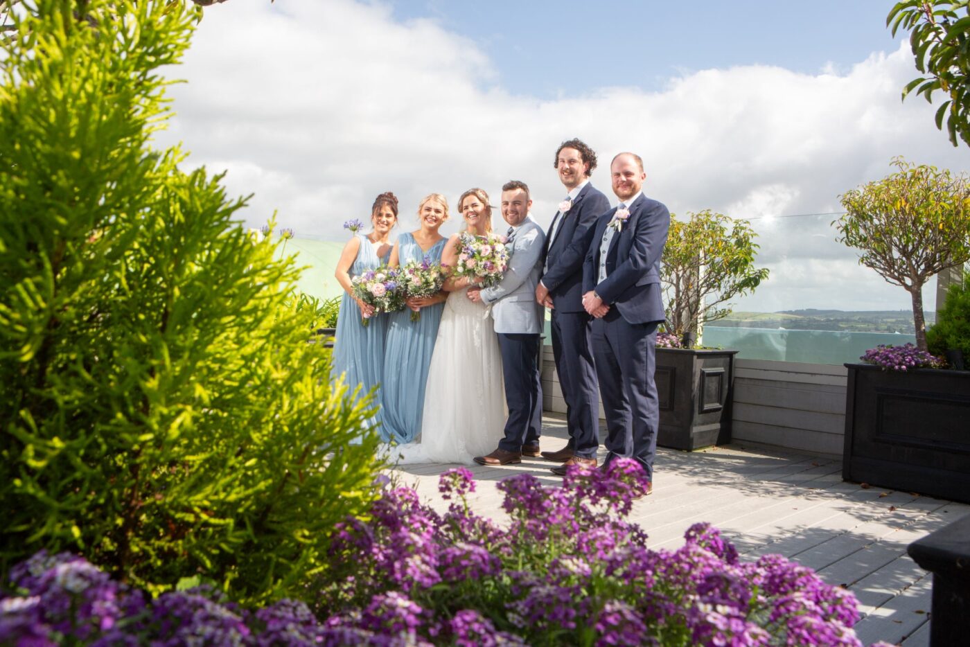 Bride and groom with bridesmaids in pale blue and groomsmen in navy on a sunny rooftop terrace framed by flowers and potted trees.