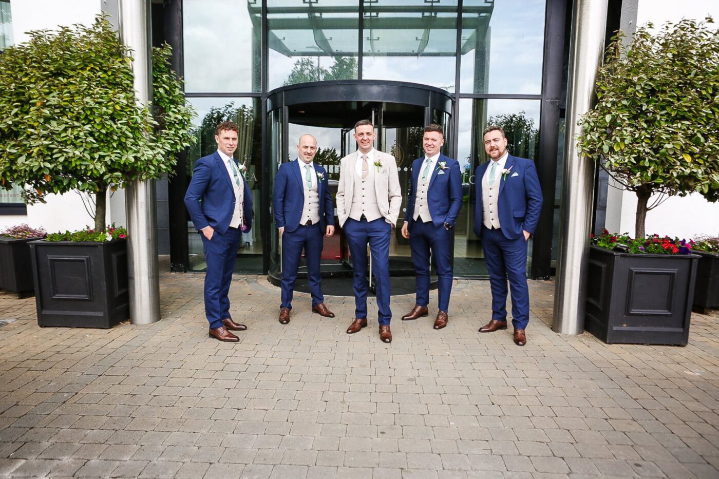 Groom in a cream jacket with four groomsmen in navy posing outside a hotel’s revolving doors between potted trees outside the loughrea hotel
