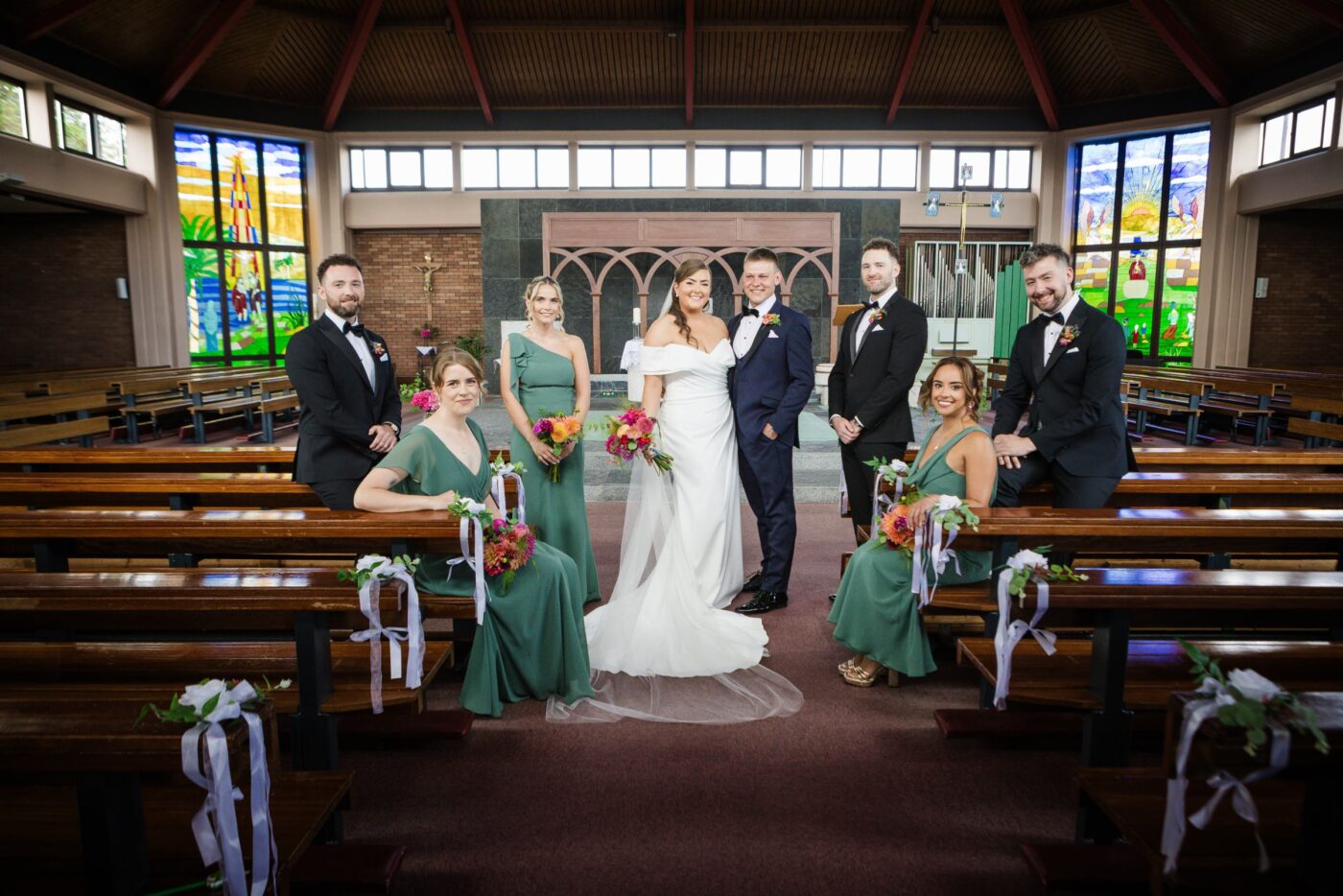 Bride and groom with bridesmaids in sage green and groomsmen in black, beneath colourful church stained-glass windows.
