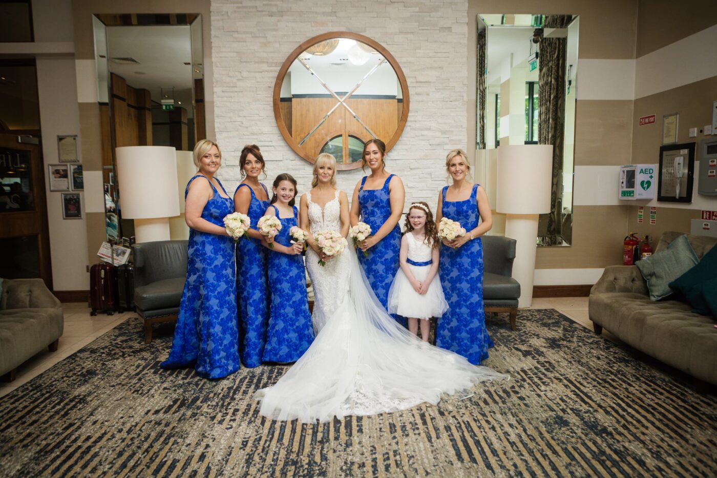 Bride with bridesmaids in blue floral dresses and a flower girl beside a hotel fireplace.