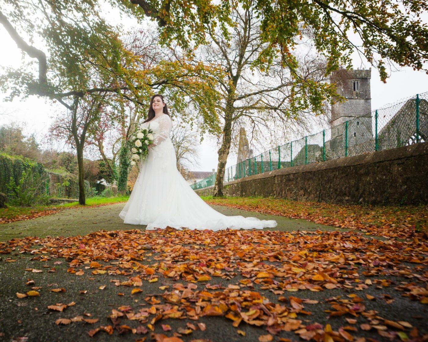 Bride holding a white bouquet beneath autumn trees, with fallen leaves and stone abbey ruins behind her.