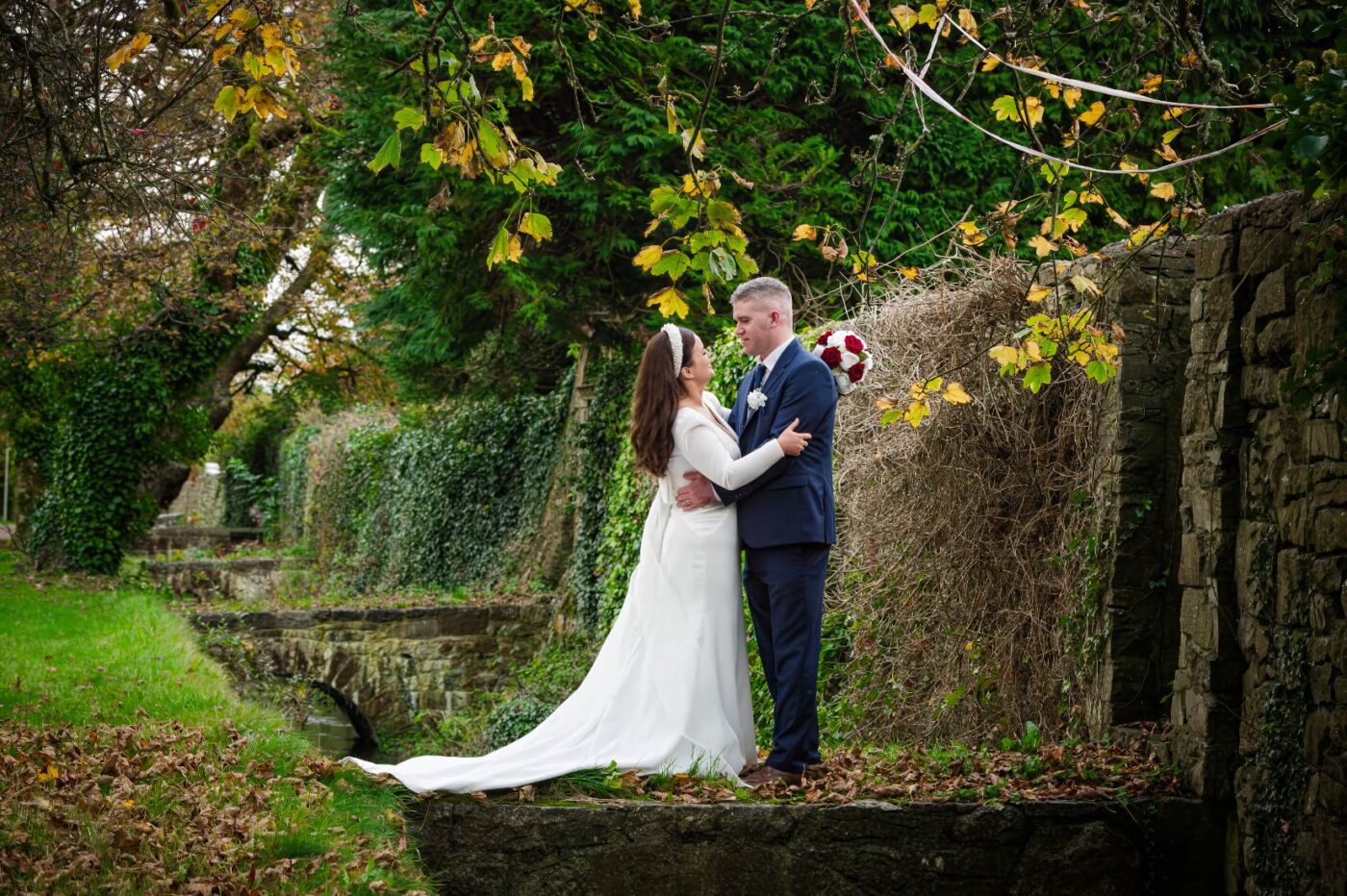 Bride and groom embracing beside an ivy-covered stone wall and stream beneath yellow autumn leaves.