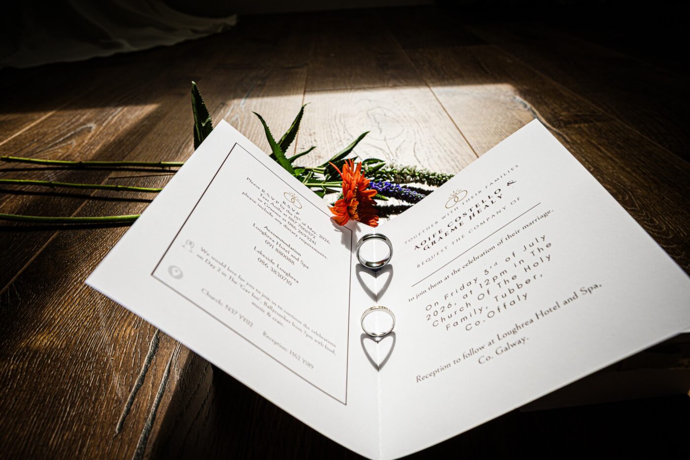 Wedding rings resting on an open invitation beside colourful flowers, with sunlight casting heart-shaped shadows.