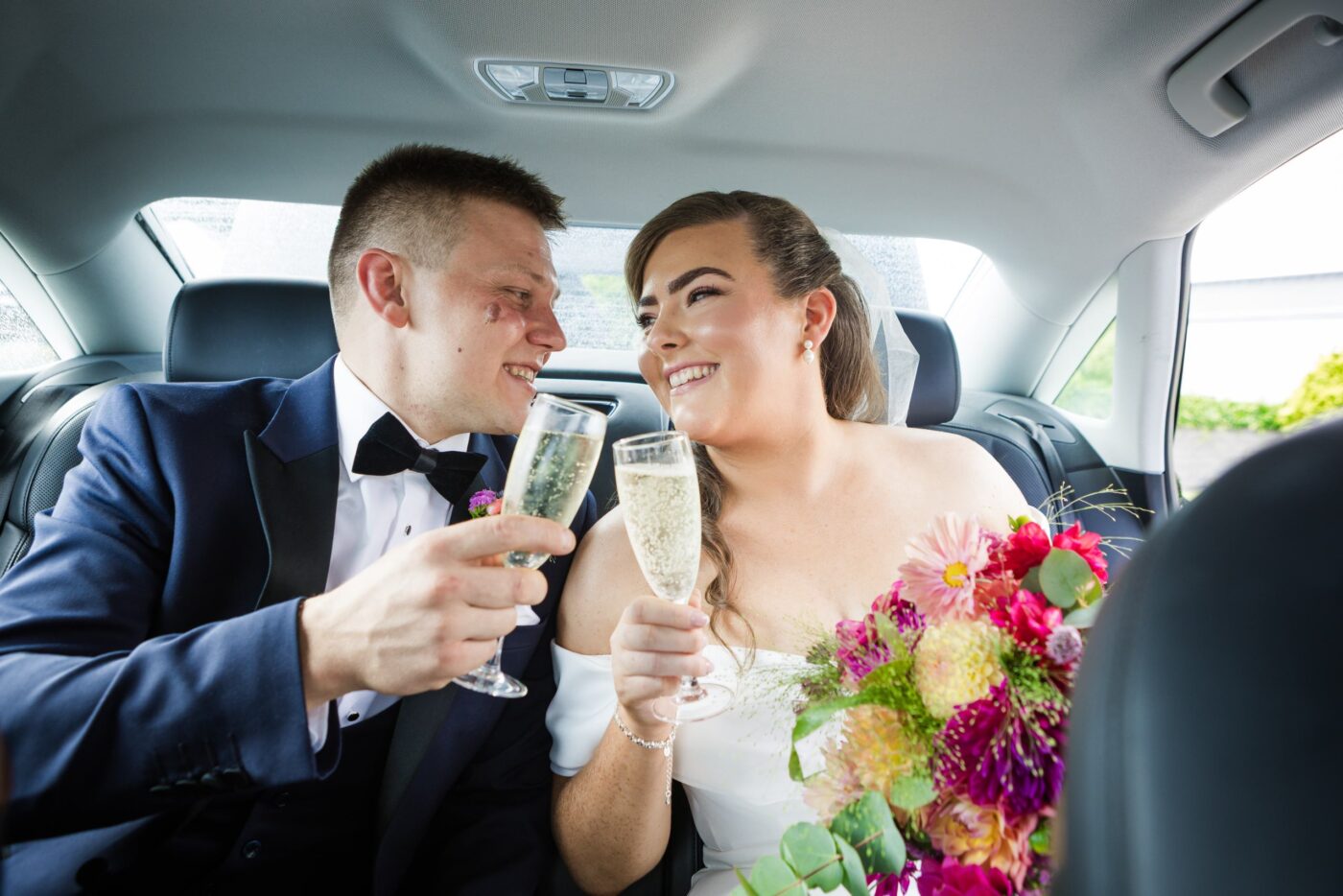 Bride and groom smiling at each other and holding champagne glasses in the back of their wedding car.