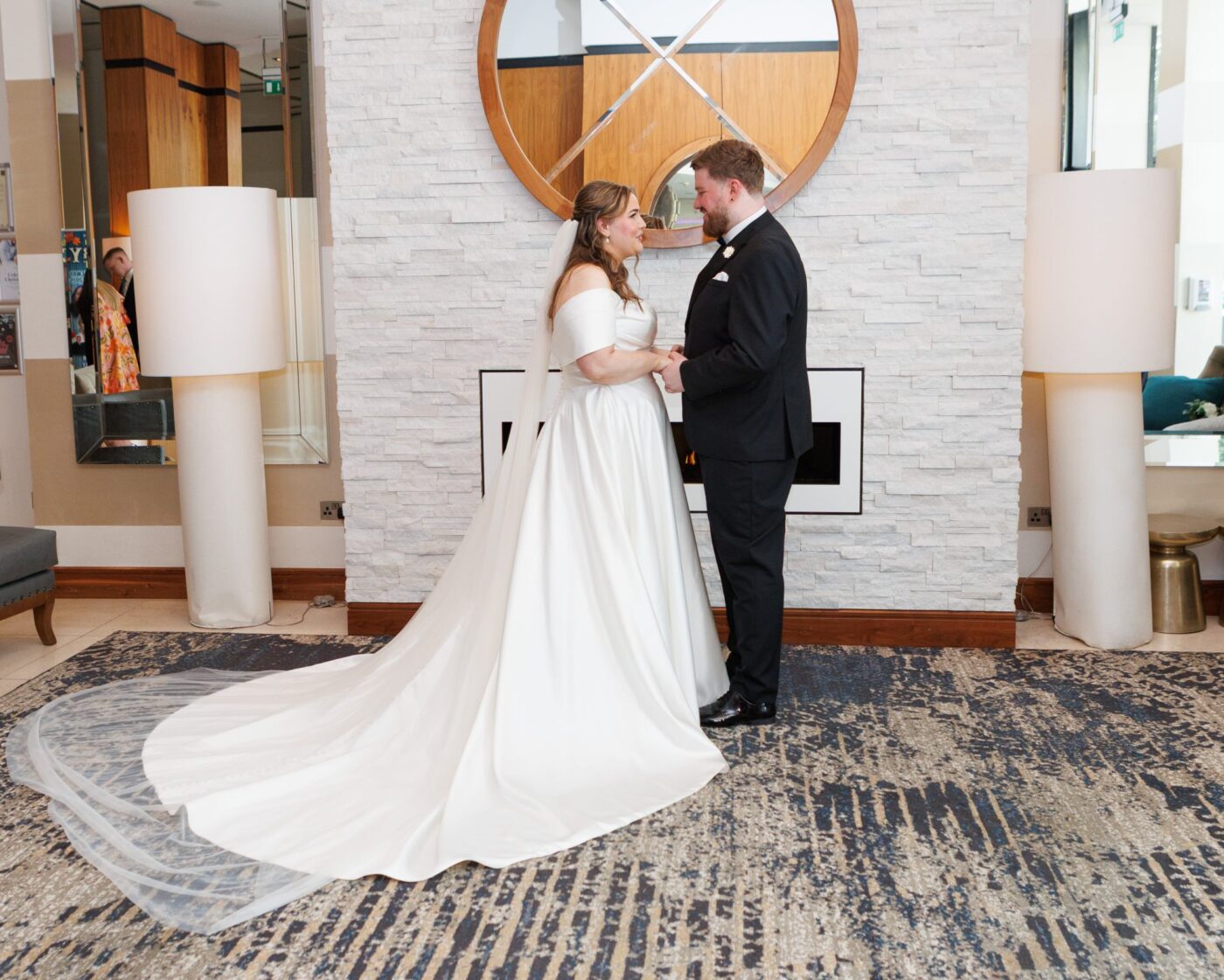 Bride and groom holding hands beside a fireplace and round mirror, with her dress train spread across the carpet.