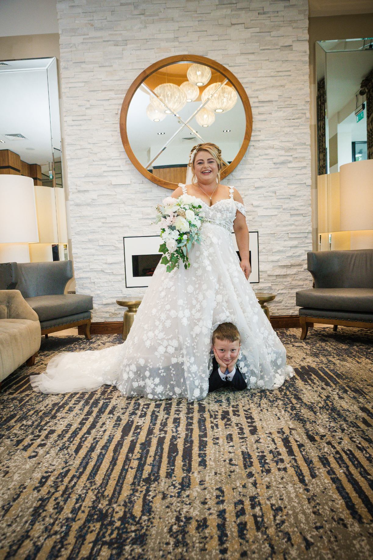 Smiling bride holding a cascading bouquet as a young boy peeks out from beneath her wedding dress.
