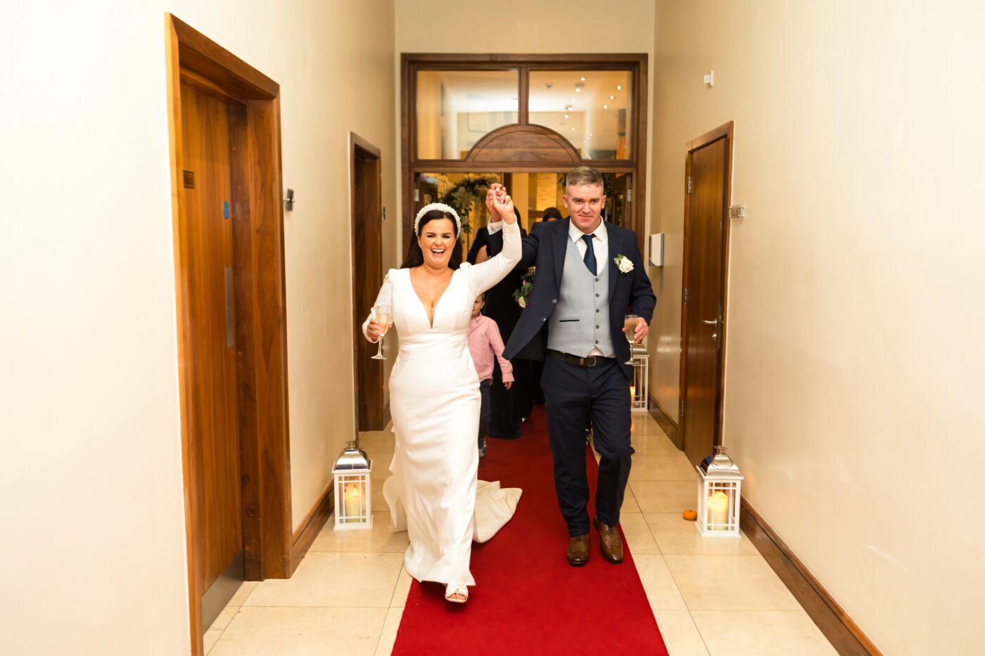 Bride and groom walking along a red carpet, raising their joined hands and holding champagne glasses.