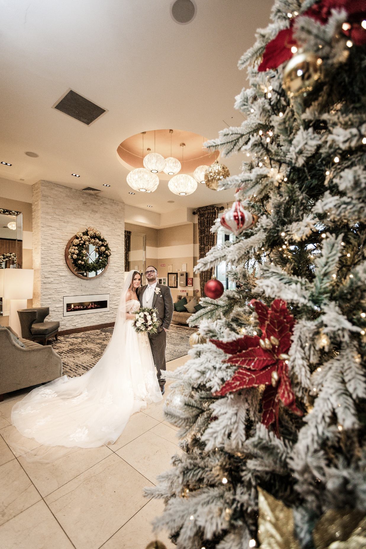 Bride and groom beside a fireplace, framed by a Christmas tree decorated with red and gold ornaments.