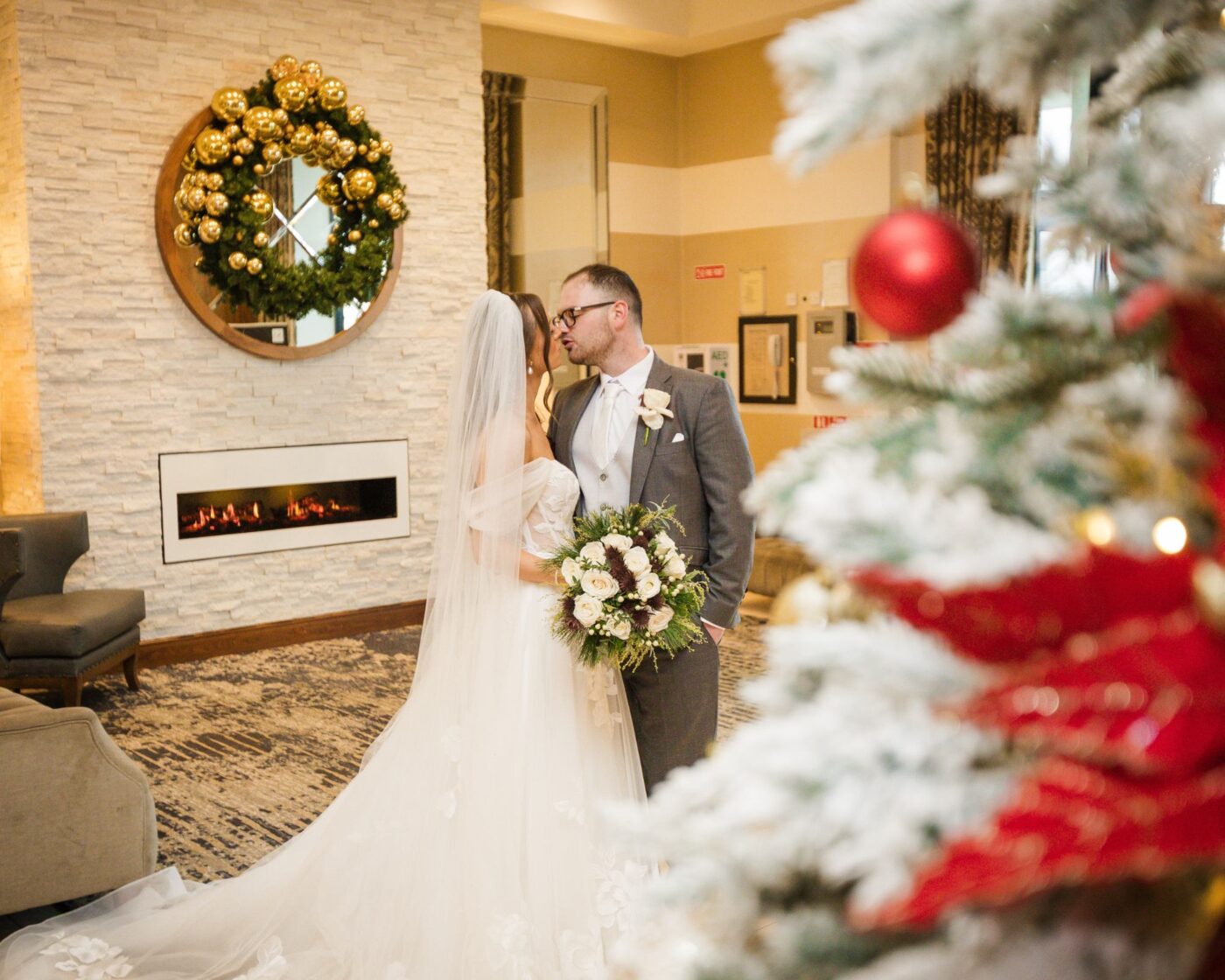 Bride and groom kissing beside a hotel fireplace, with a decorated Christmas tree in the foreground.