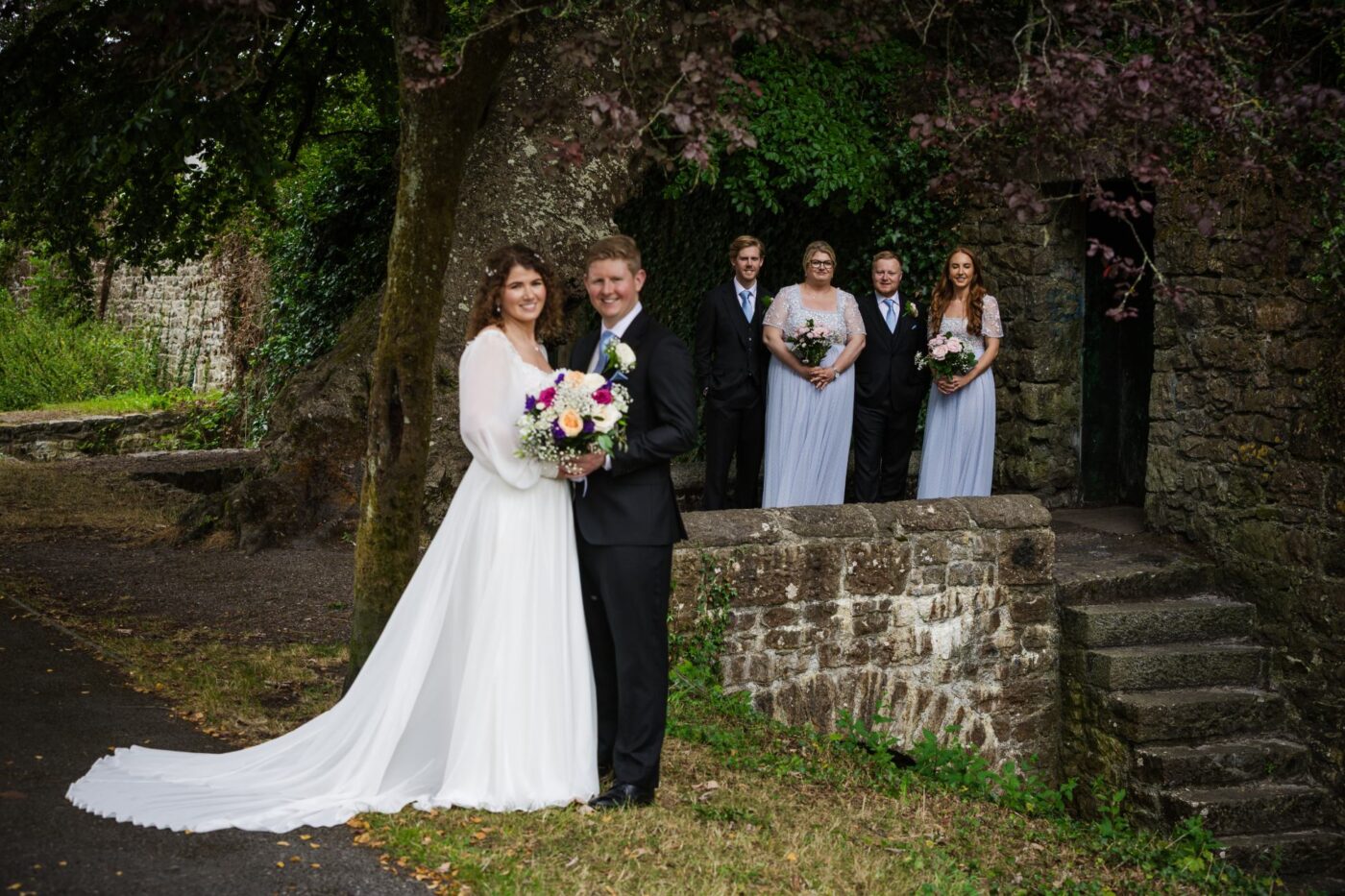Bride and groom standing beside a tree, with bridesmaids and groomsmen behind a stone wall at ivy-covered ruins.
