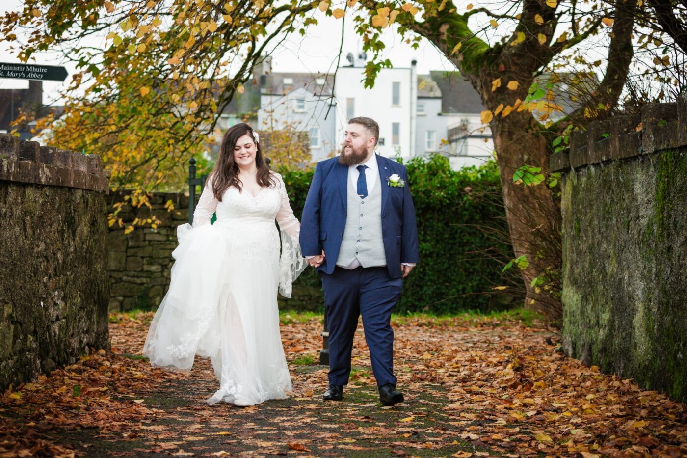 Bride and groom holding hands on a leaf-covered path between stone walls beneath autumn trees.