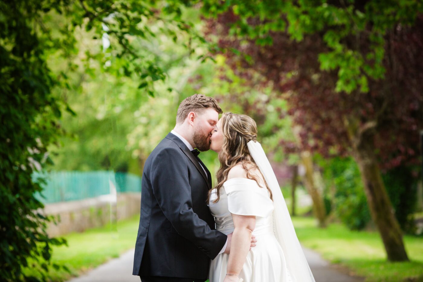 Bride and groom kissing on a tree-lined garden path surrounded by green foliage.