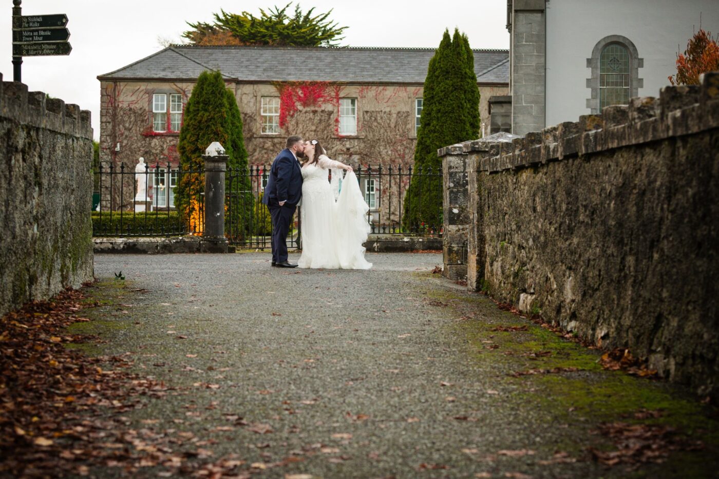 Bride and groom kissing on a stone-walled path, with an ivy-covered building behind them.