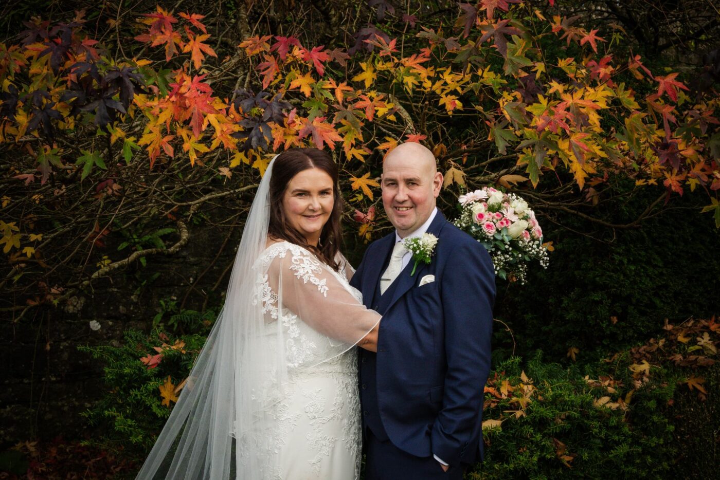 Bride and groom smiling beneath red and orange autumn leaves, with a pink and white bouquet over his shoulder.