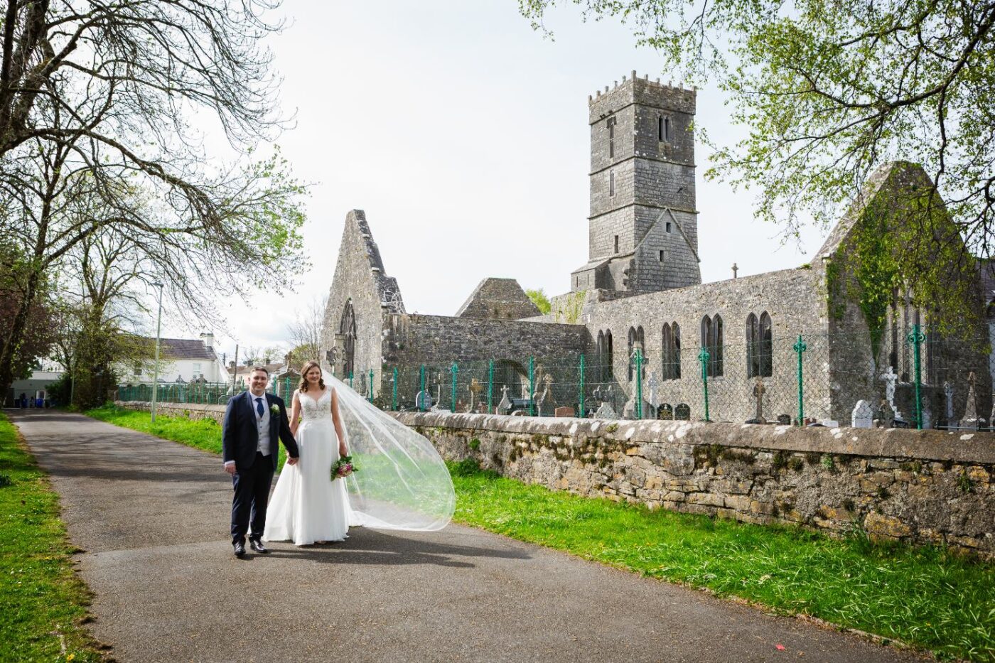 Bride and groom walking hand in hand beside stone abbey ruins, with her veil blowing behind her.