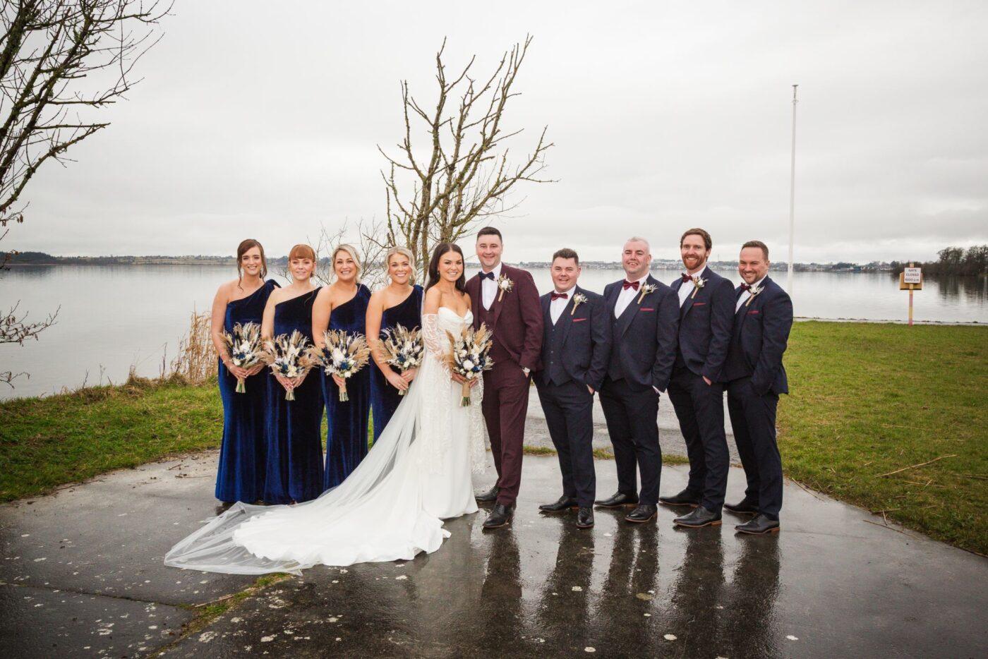 Bride and groom with bridesmaids in navy velvet and groomsmen in navy suits, reflected on a wet lakeside path.