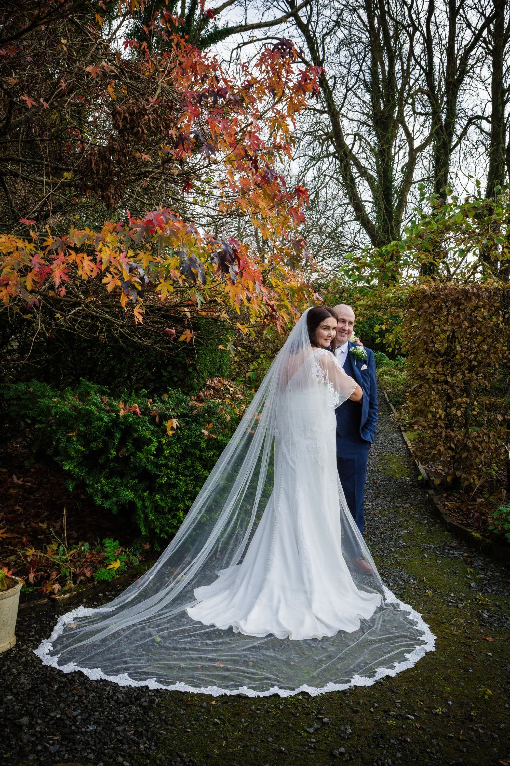 Bride looking back towards the camera beside the groom, with her lace-edged veil spread across an autumn garden path.