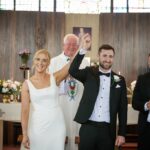 Áine and Rory raising their joined hands as Father Bohan smiles and their bridal party applauds.