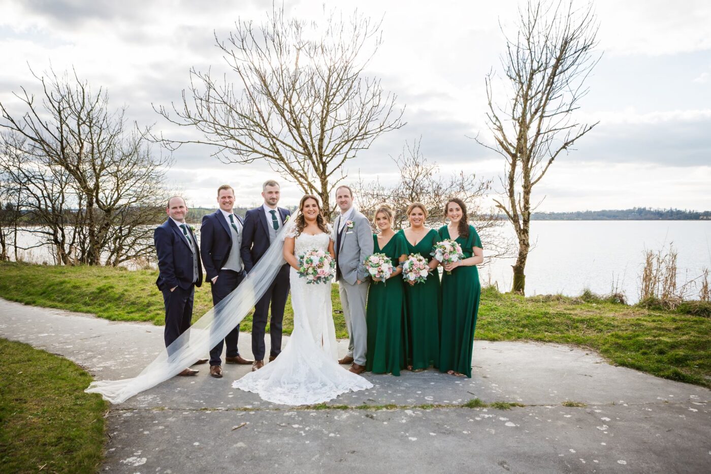 Bride and groom beside a lake with bridesmaids in emerald green and groomsmen in navy, with her veil flowing to the side.