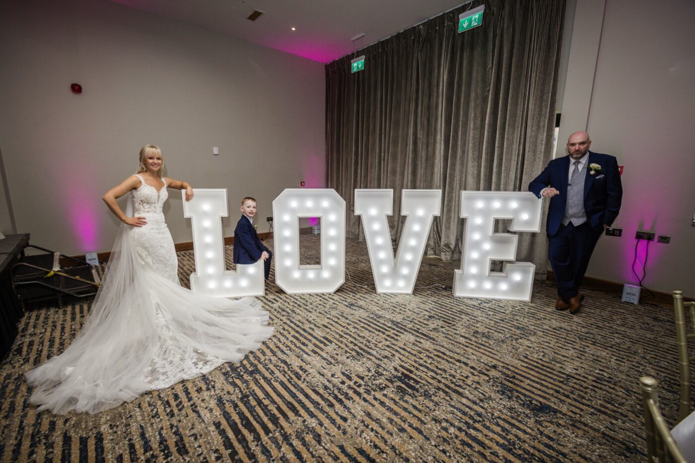 Bride and groom with a young boy beside illuminated LOVE letters in the wedding reception room at Loughrea Hotel & Spa.