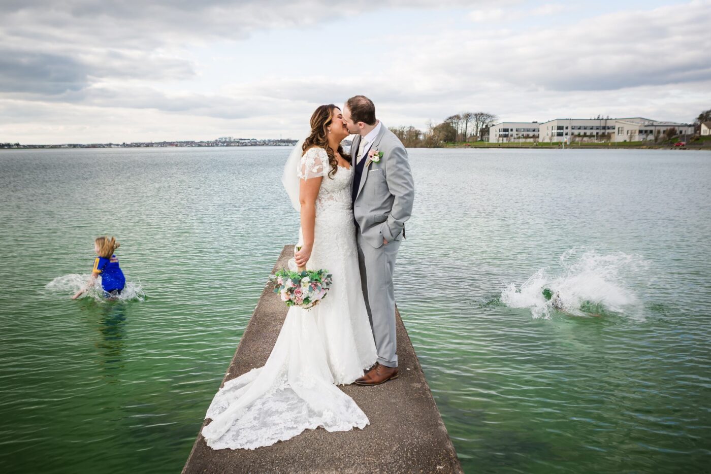 Bride and groom kissing on a pier at Loughrea Lake as swimmers splash into the water on either side.